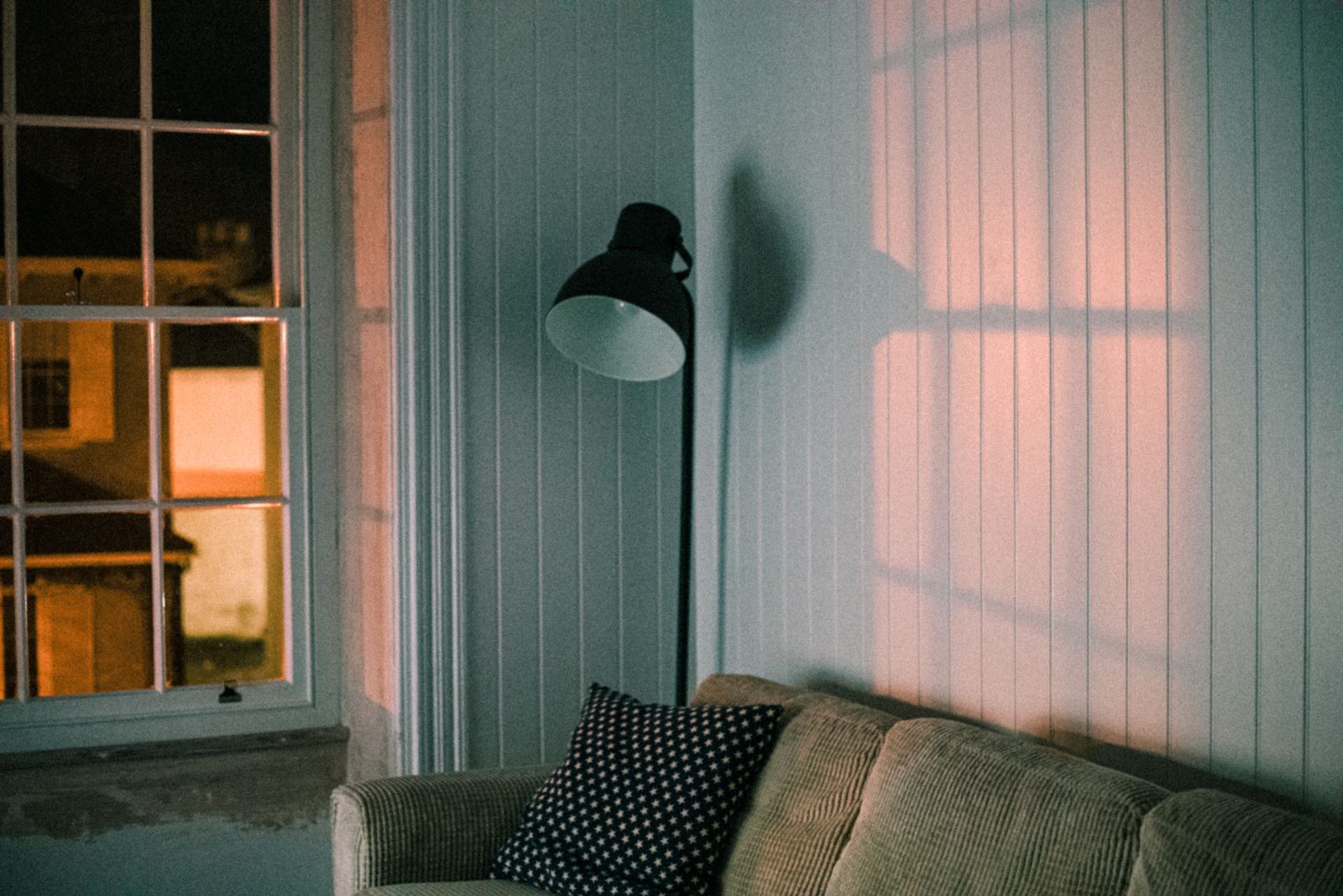 Un salon le soir, un lampadaire noir allumé à côté d'un canapé gris près d'une fenêtre