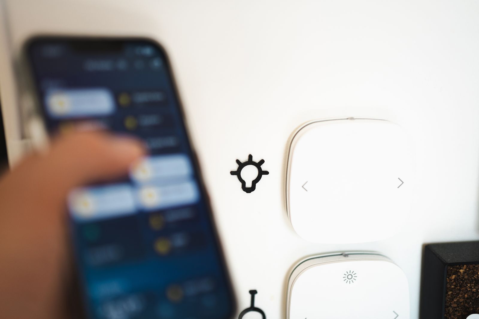 Une main tient un smartphone ouvert sur une application de maison connectée, devant des interrupteurs muraux connectés